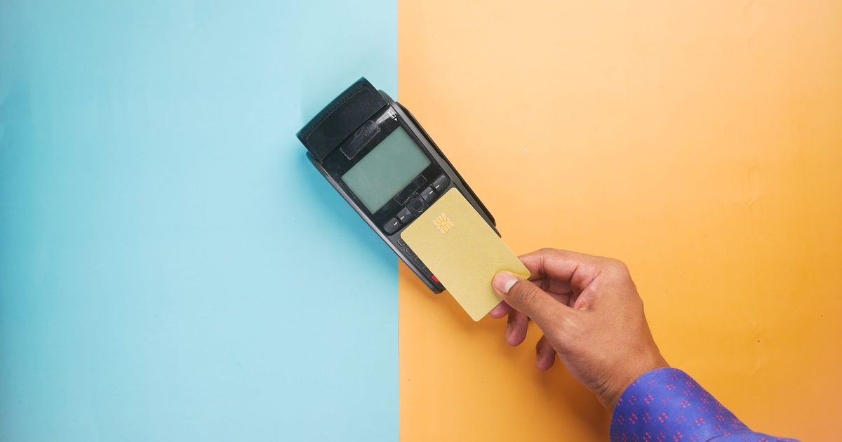 A hand holding a gold payment card against a card terminal on a split blue and orange background