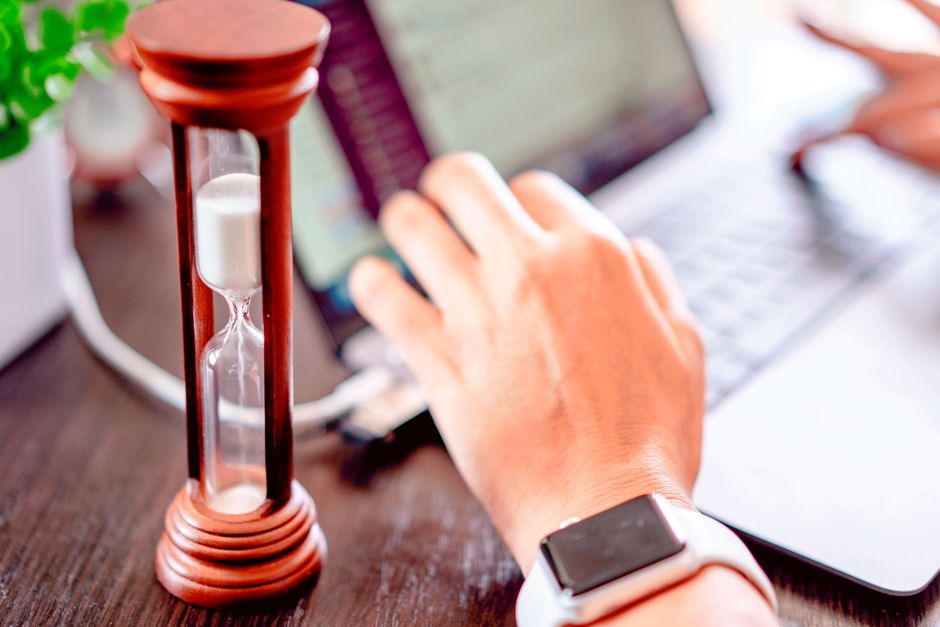 A wooden hourglass beside a hand typing on a laptop, with a smartwatch on the wrist